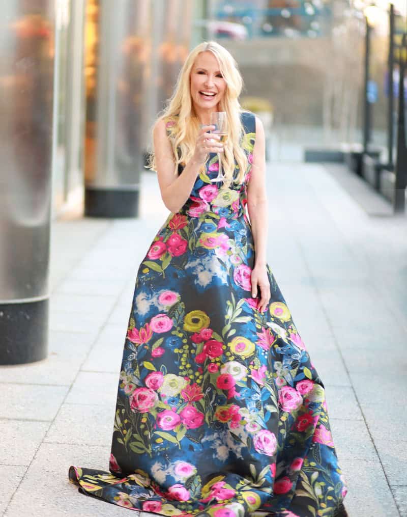 Elegant woman wearing a vibrant floral maxi dress, holding a glass of wine, smiling outdoors on a sunny day, showcasing fashion, style, and personality.