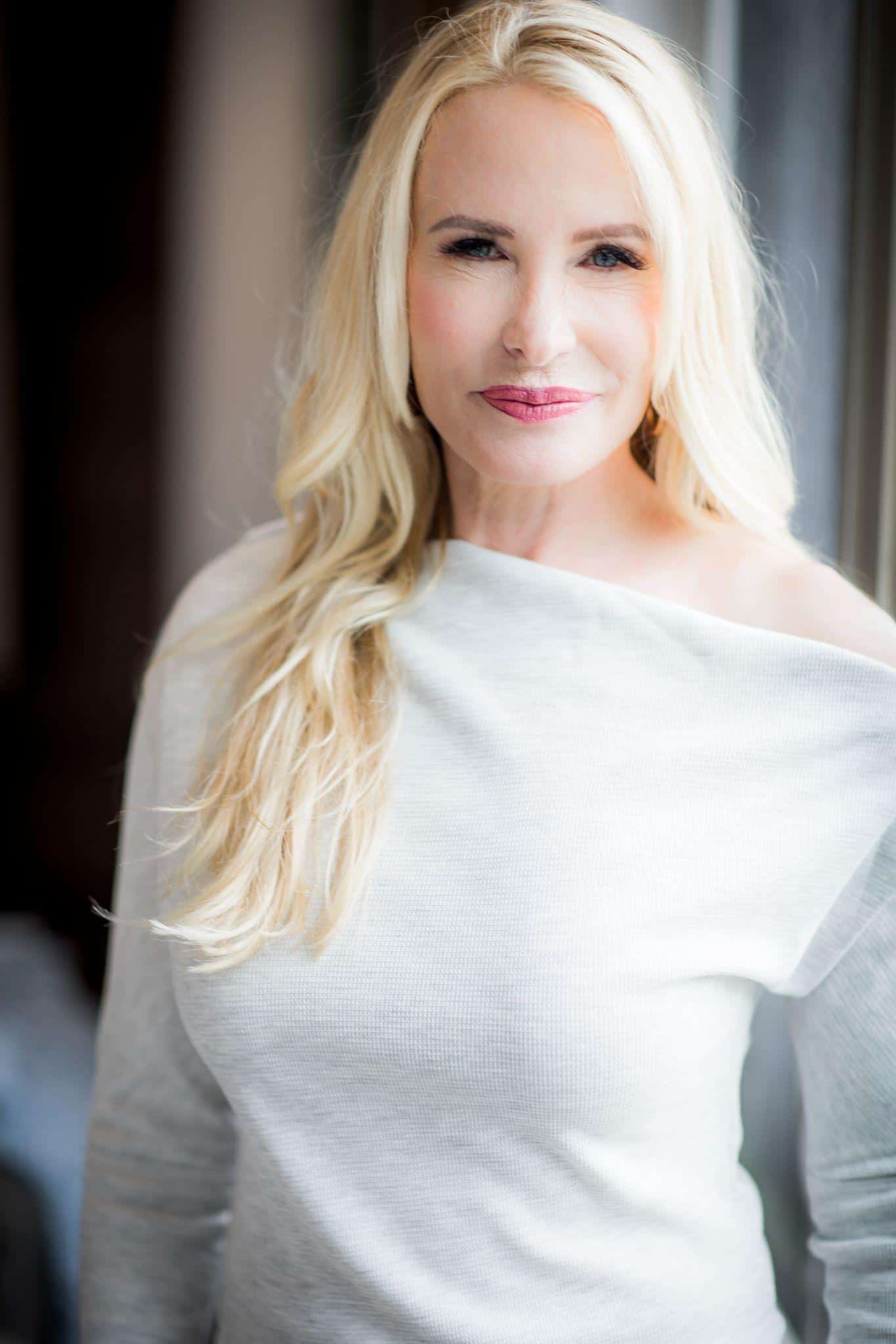 Elegant blonde woman with soft makeup looking confidently at the camera, wearing a light-colored off-shoulder top indoors, showcasing her beauty and style.