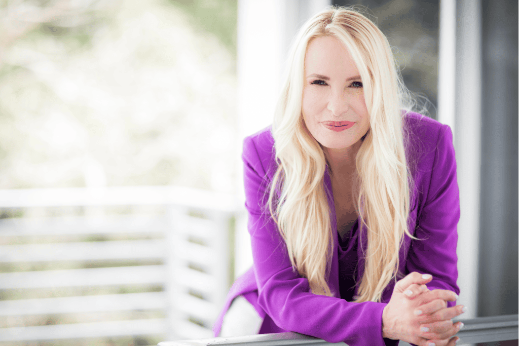 Elegant blonde woman in purple blazer leaning on balcony railing, outdoor natural light, professional business portrait, confident and approachable.